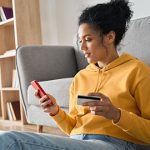 Woman with yellow sweatshirt in home sitting against couch looking at phone and holding a bank card.